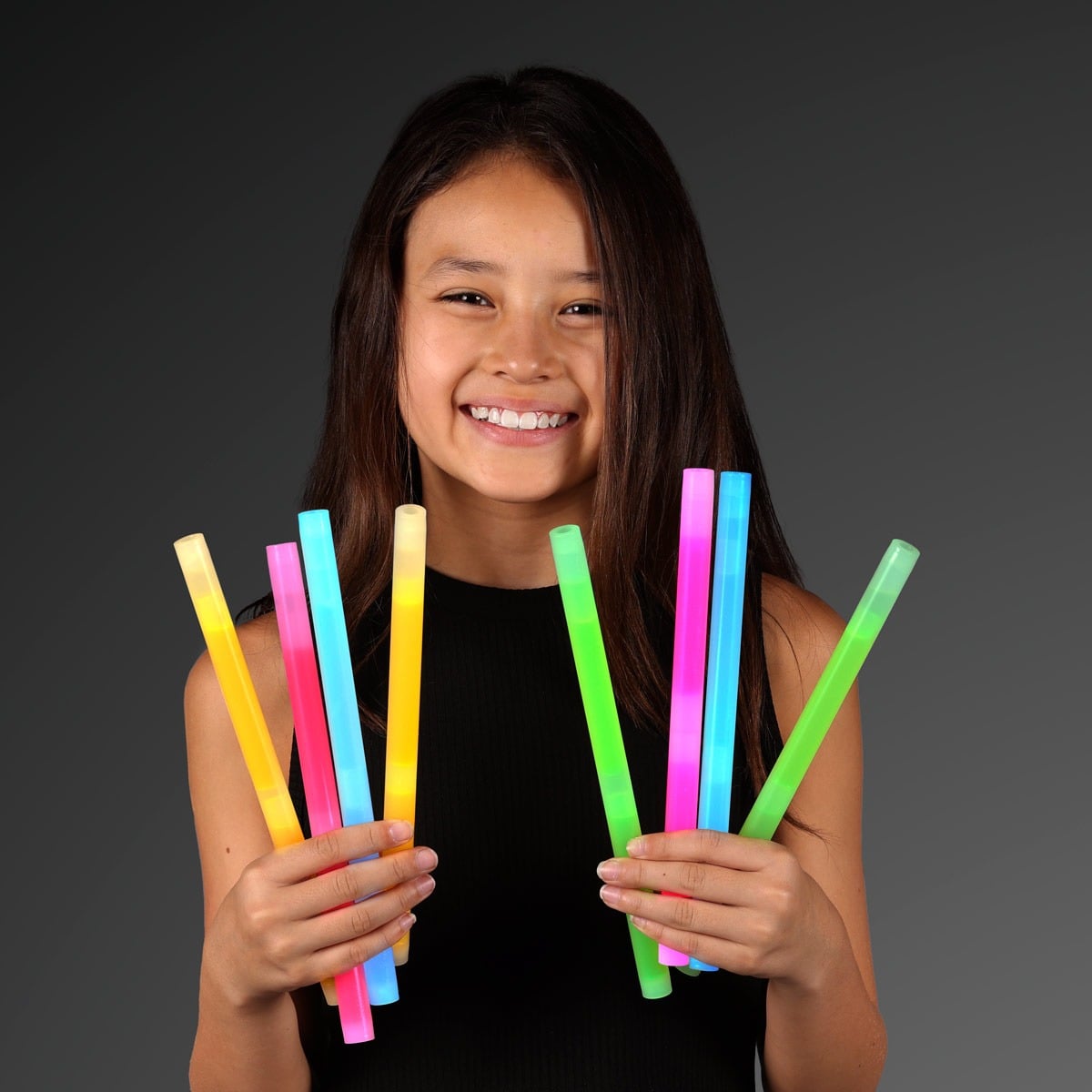 A smiling girl with long brown hair, in a sleeveless black top, holds several colorful 9.4 Blow Stick Wands in both hands against a dark gray background.